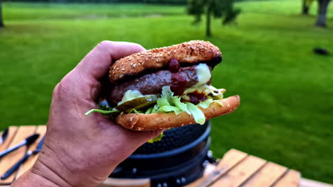 hand holding mouthwatering hamburger, eating out at park at sunset. pov, tilt-up shot