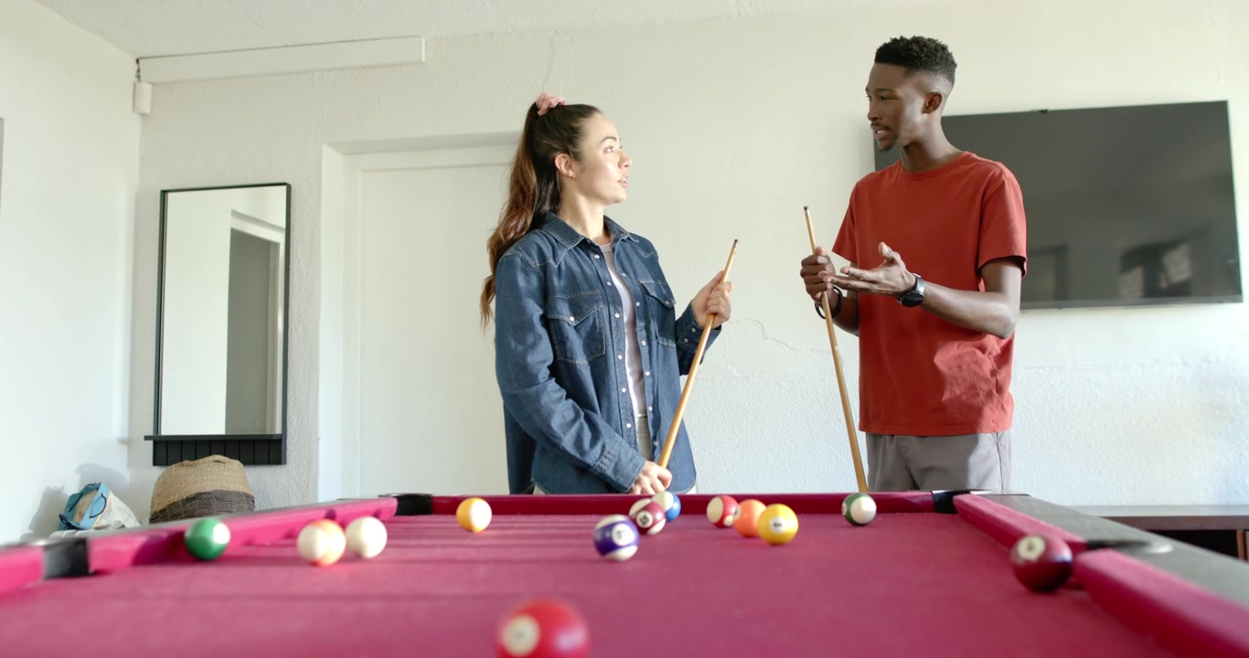 Diverse couple behind red-felt table, man cupping in red tee explaining shot holding cue near balls