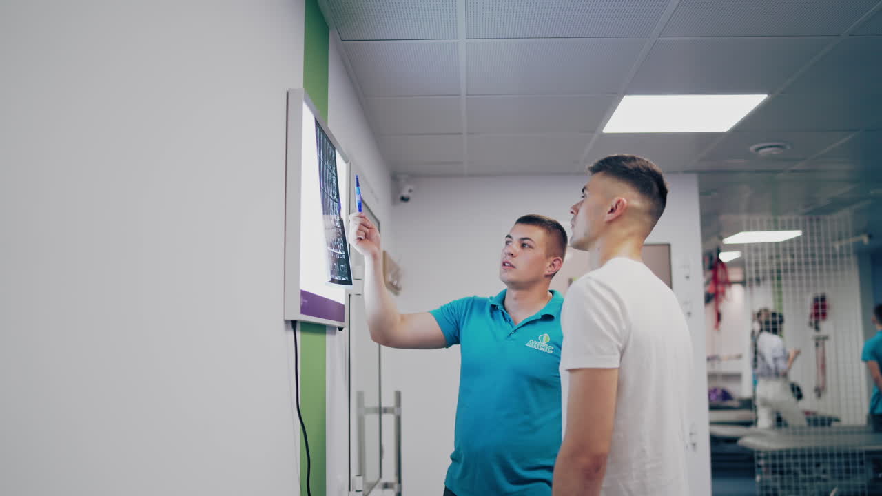Young doctor shows the problem areas of the back on the X-ray to the patient. Therapist man talks to a patient and shows spine x-ray on a white board in clinic.