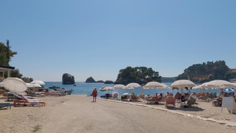 Toma-Panorámica-Lenta-De-Turistas-Relajándose-Bajo-Sombrillas-En-La-Impresionante-Playa-De-Parga,-Grecia