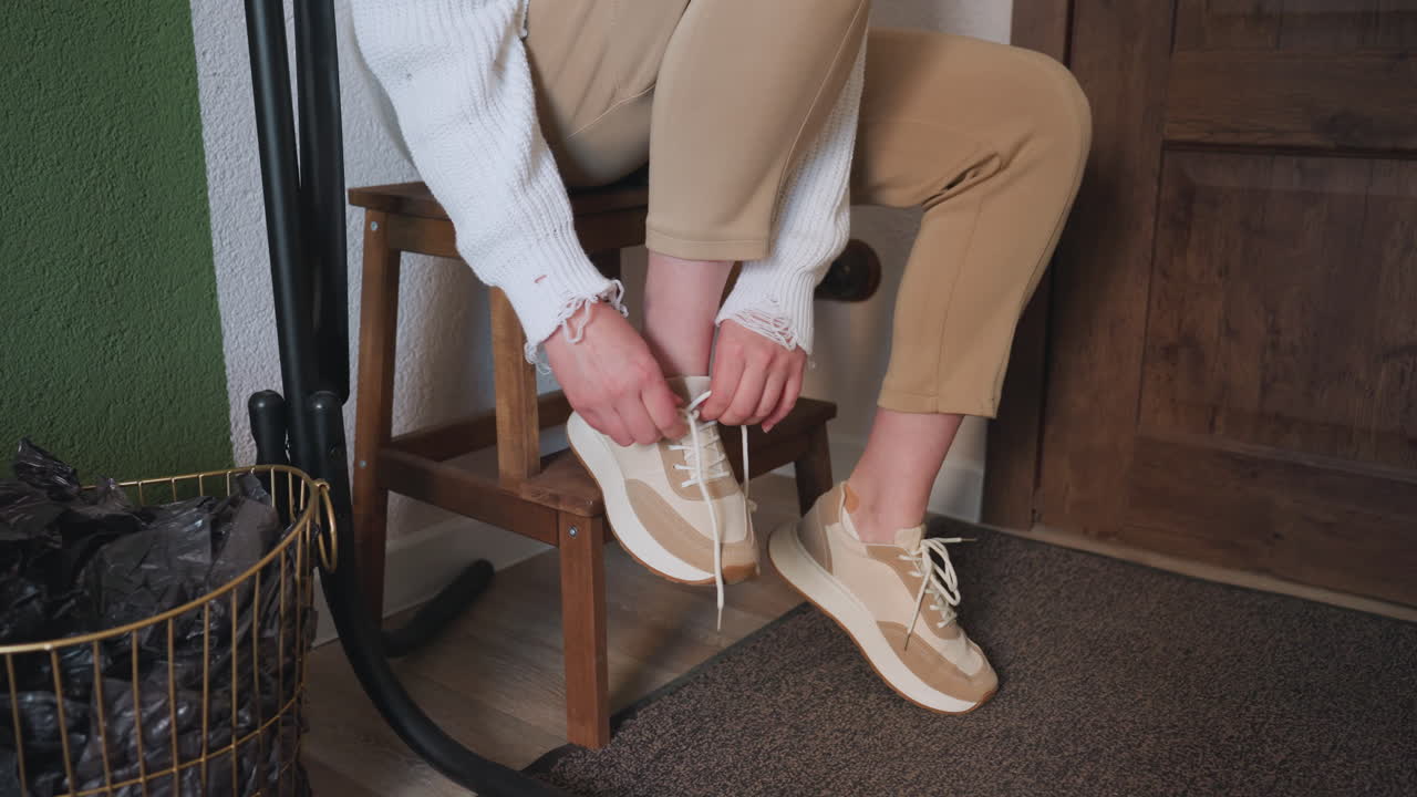 Leg view of client seated on wooden bench removing beige canvas shoes before session, with neutral pants, white sweater, green wall, coat stand, and indoor decor creating calm
