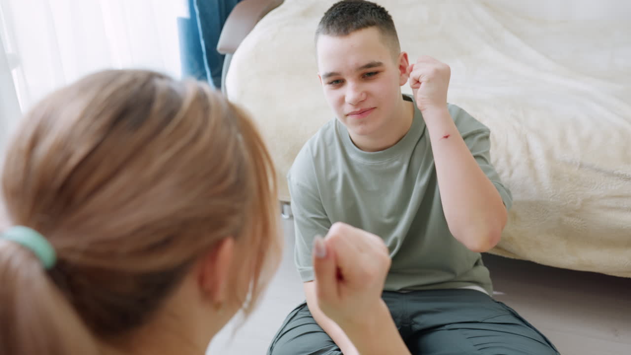 Mother and son sitting cross-legged on floor near window laughing and playing hand trick game, enjoying quality bonding time in bright modern room with indoor plant