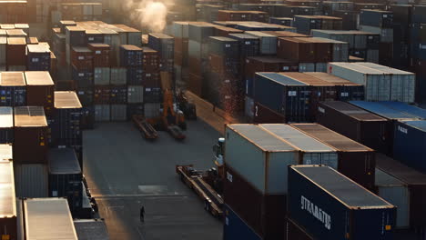 Aerial view of a bustling container port at sunrise or sunset with stacked shipping containers and a lone worker