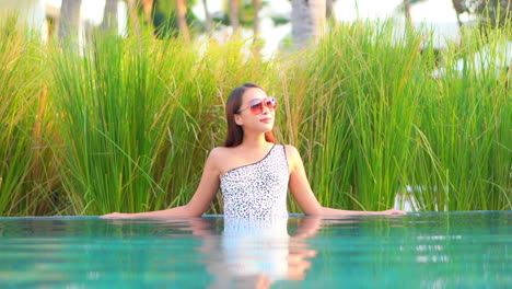 Mujer-Coreana-De-Moda-En-Monokini-Manchado-De-Un-Hombro-Relajándose-En-El-Agua-De-La-Piscina-Apoyada-En-El-Borde-De-La-Piscina-En-El-Salón-Del-Hotel-Tropical-En-Malasia,-Cámara-Lenta-Estática