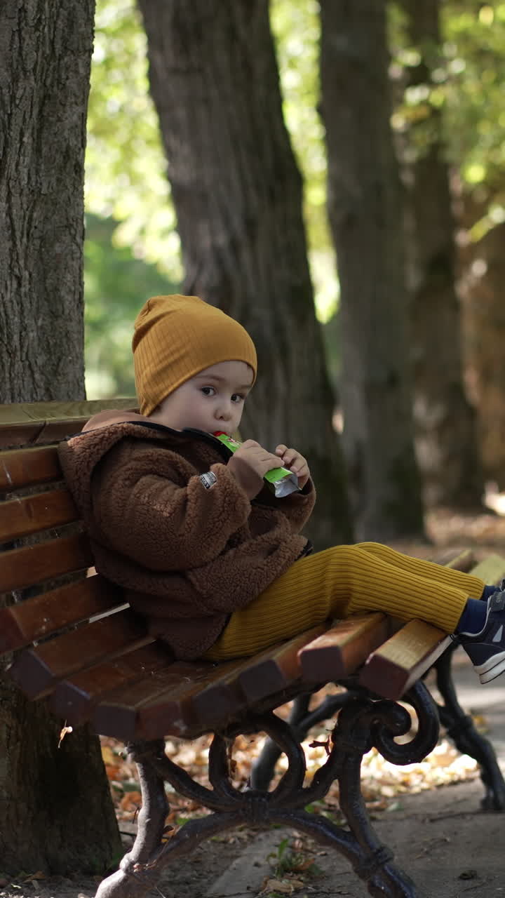 Cute baby boy sits peacefully on the bench eating. Cute kid wearing warm clothes having rest during a walk in the park. Vertical video