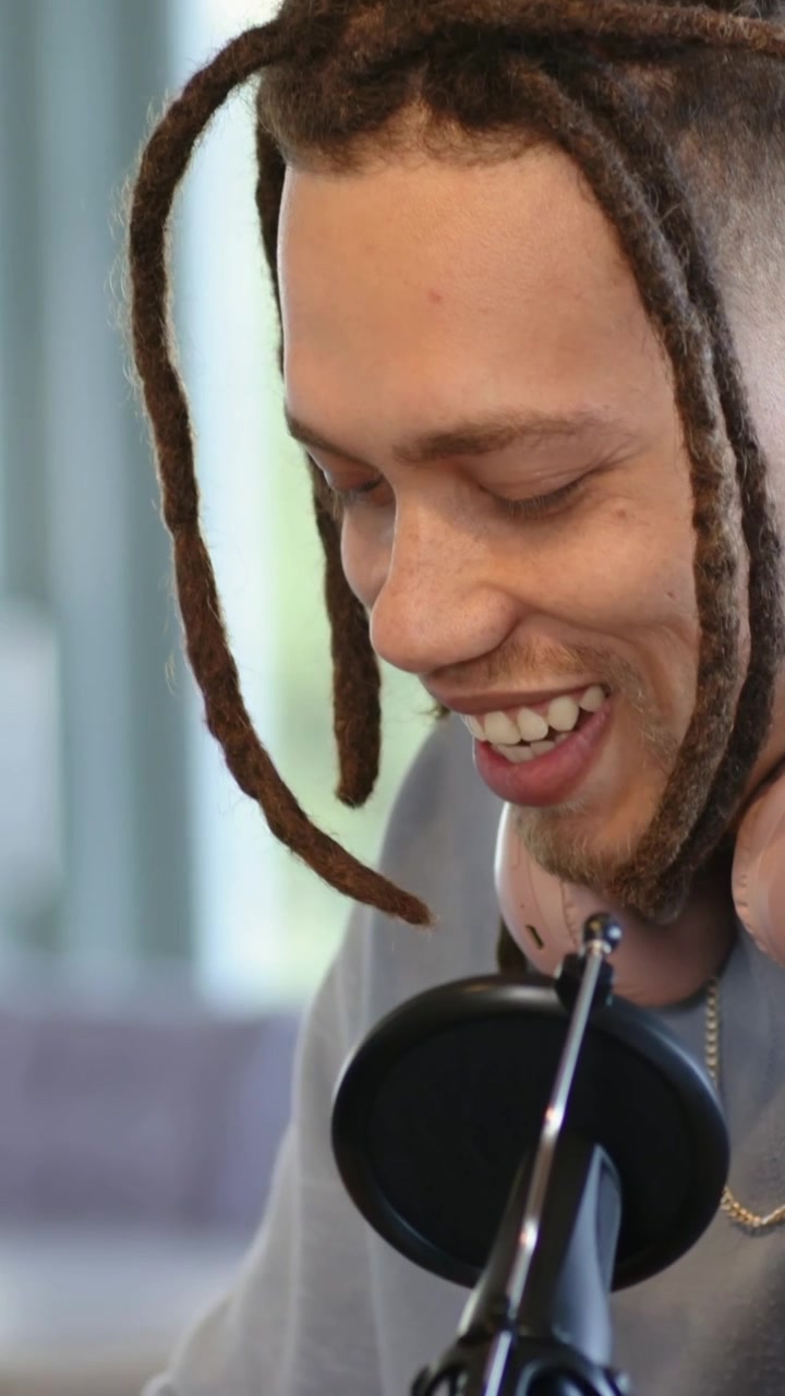 Vertikal video: Värd sträcker sig, justerar mikrofon och flyttar rosa hörlurar i hemmastudio för podcast.