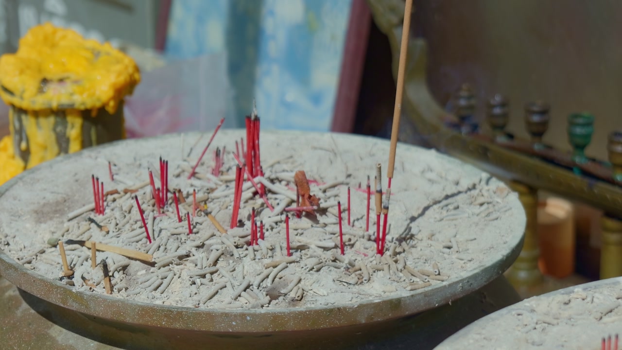Cinematic close-up of traditional red incense sticks burning in a bowl of ash. Wisps of smoke rise at a shrine in Wat Nai Harn temple, Phuket, Thailand. High-quality 4K religious footage.