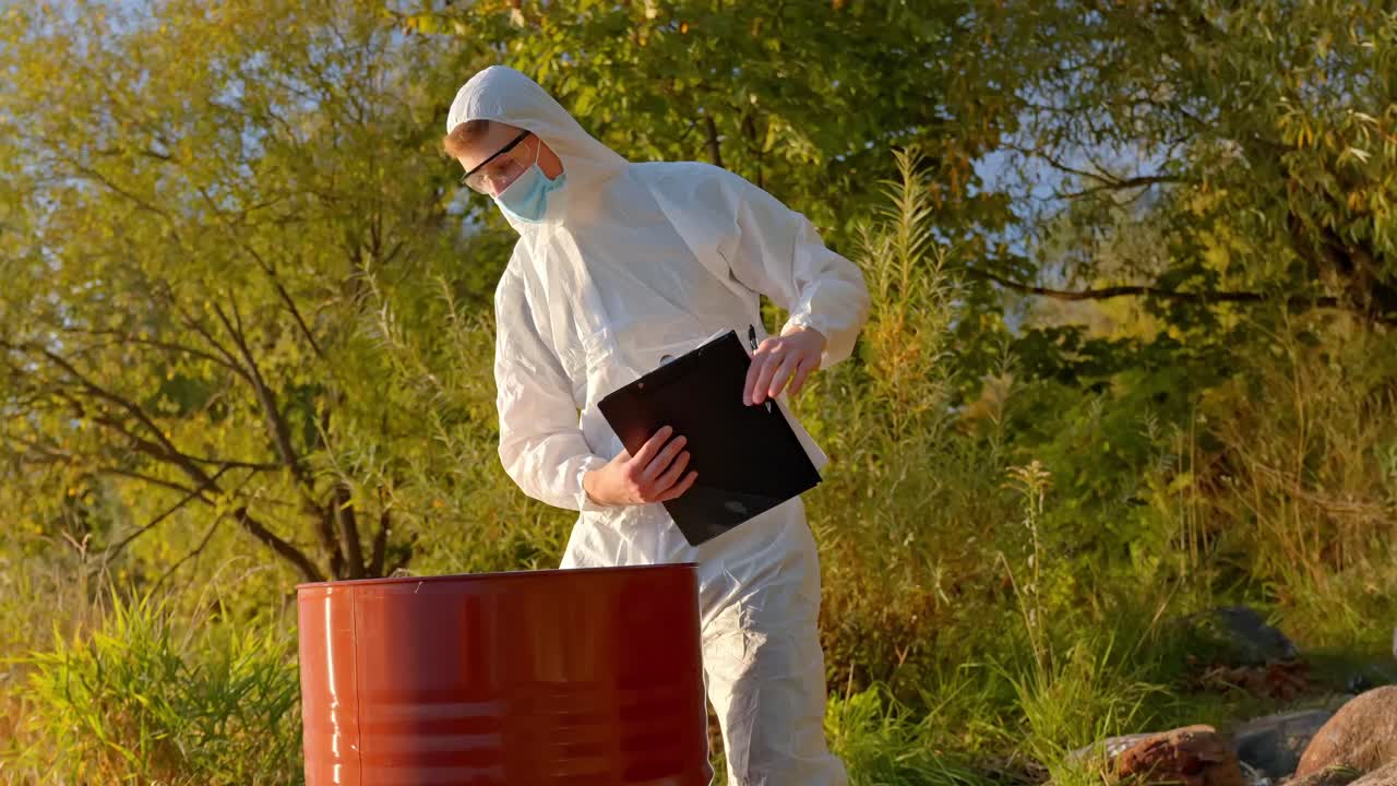 Scientist in white coveralls bends over red barrel to inspect water or leakage near leafy shoreline
