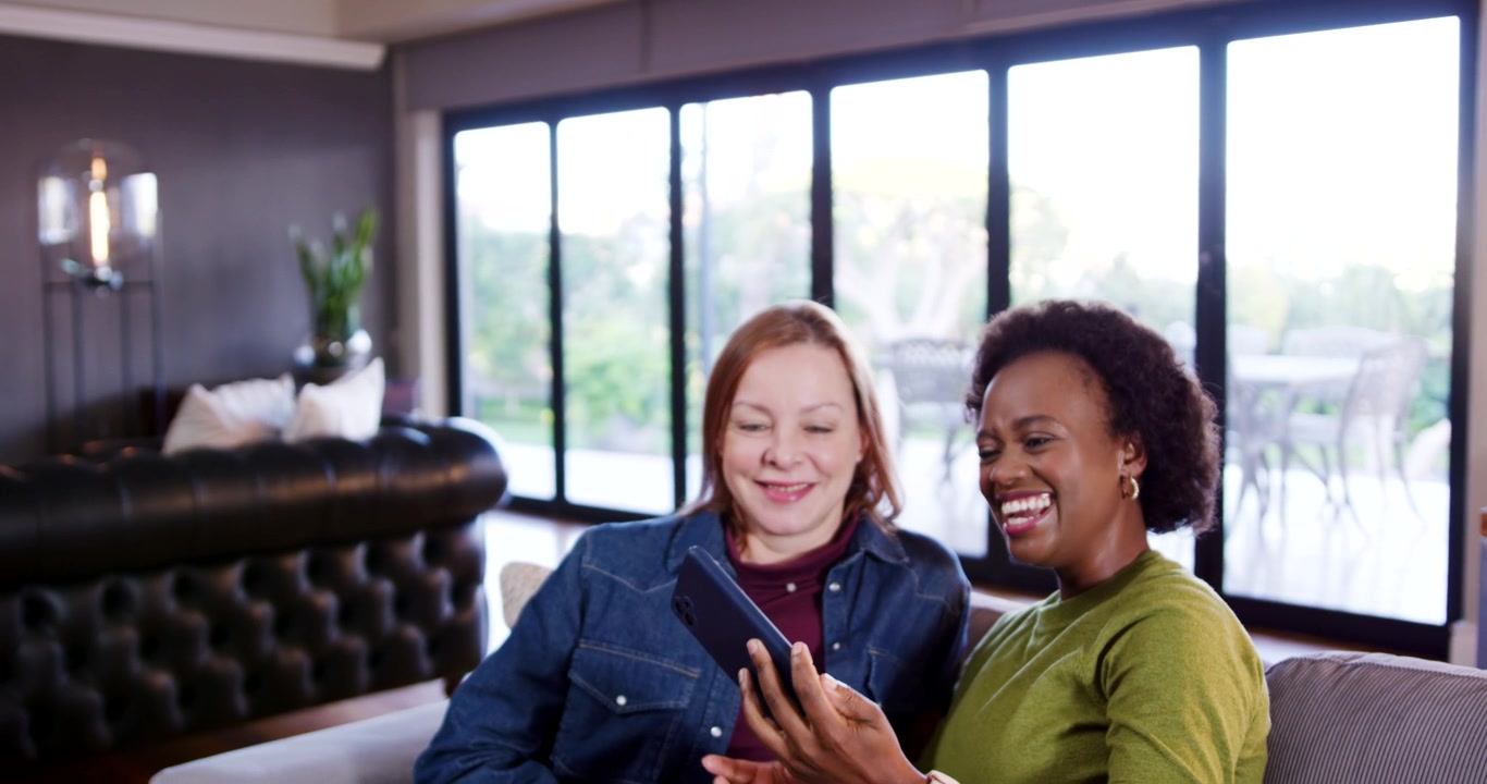 Diverse female friends raising smartphone for selfie, leaning on couch and reviewing phone with mug