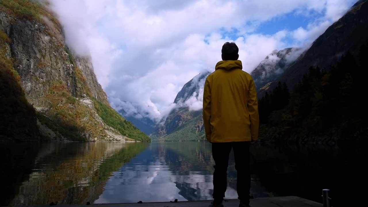 hombre con un impermeable amarillo parado frente a un fiordo cristalino de montañas imponentes
