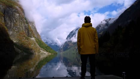 man in a yellow raincoat standing in front of a crystal clear fjord of towering mountains