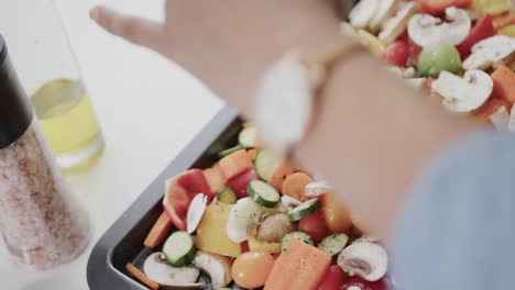 hands of biracial woman seasoning chopped vegetables on baking tray in kitchen, slow motion