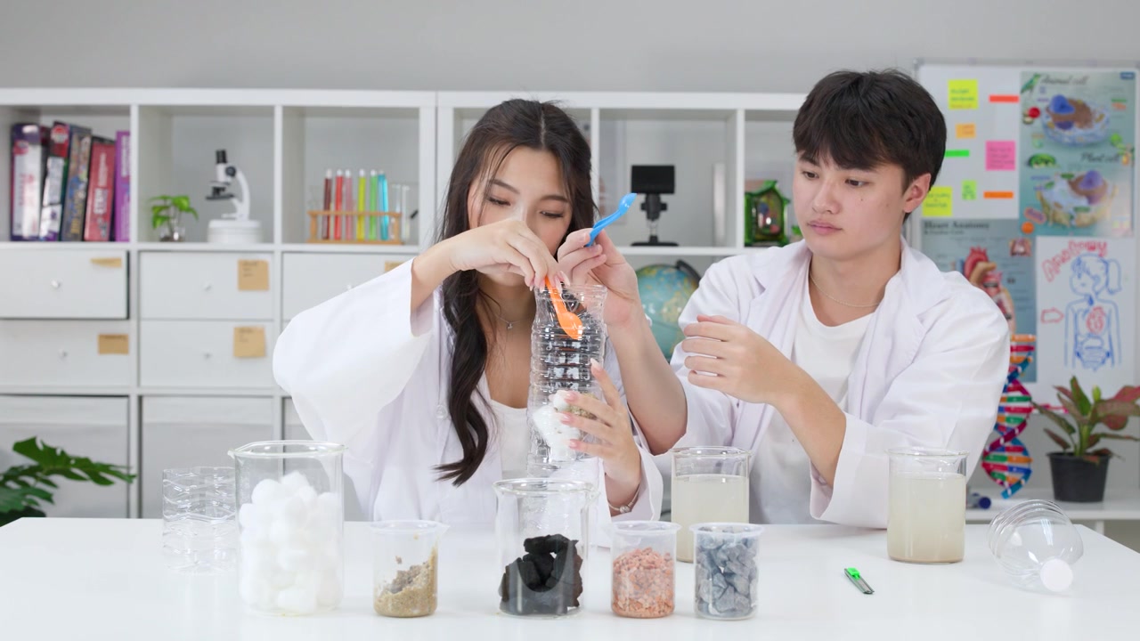 白衣を着た2人の学生が、ペットボトルと砂を使ってDIY浄水器を作っている