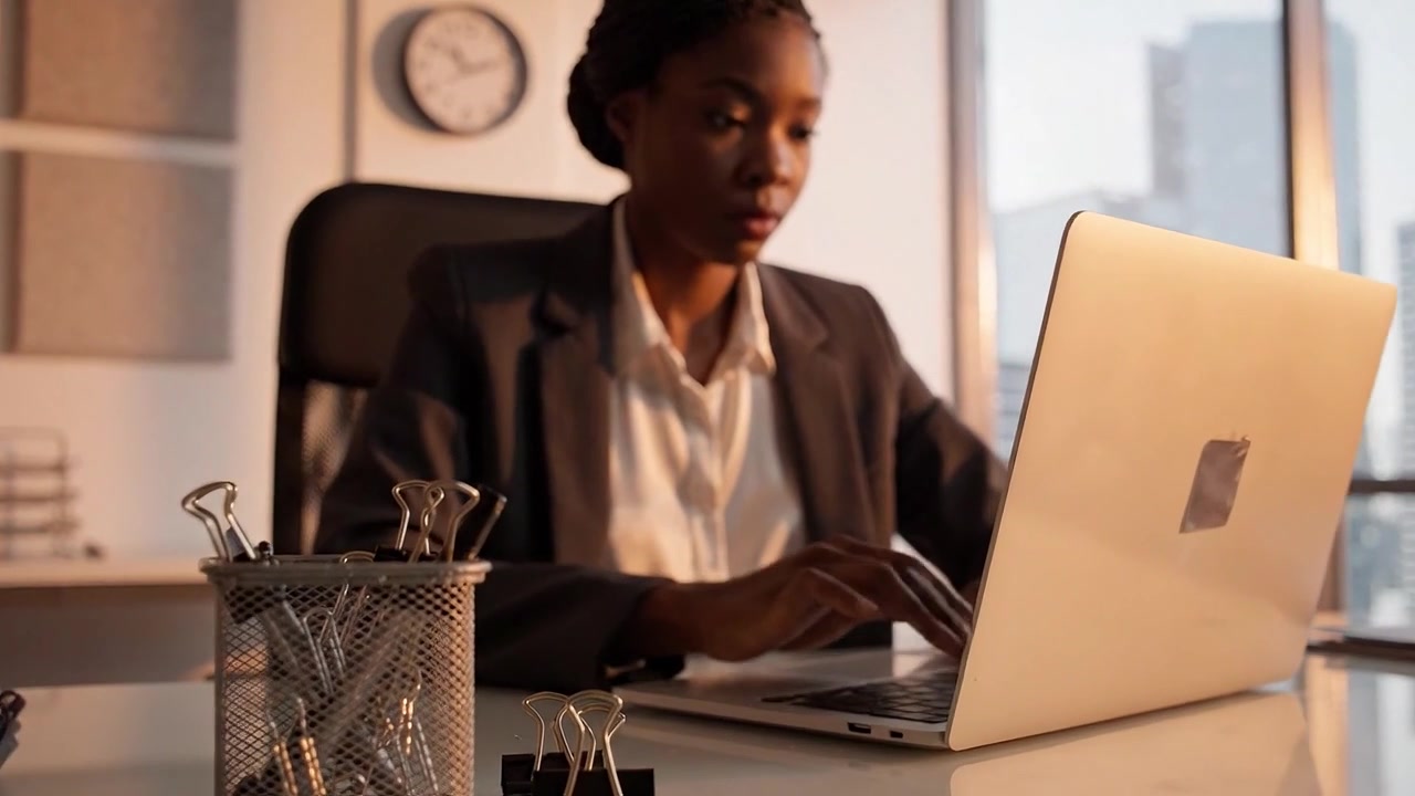 Focused Professional Working on Laptop in Modern Office Setting, Engaged in Task with Cityscape View Through Window, Clock Indicating Time Management