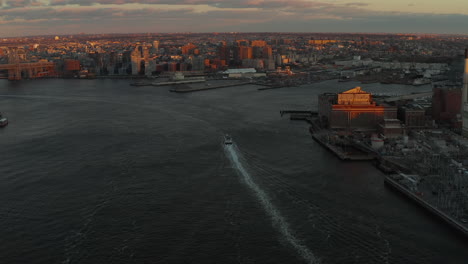 Forwards-tracking-of-boat-sailing-on-wide-river-in-city.-Tilt-up-reveal-Naval-yard-and-urban-neighbourhood-at-sunset.-Brooklyn,-New-York-City,-USA