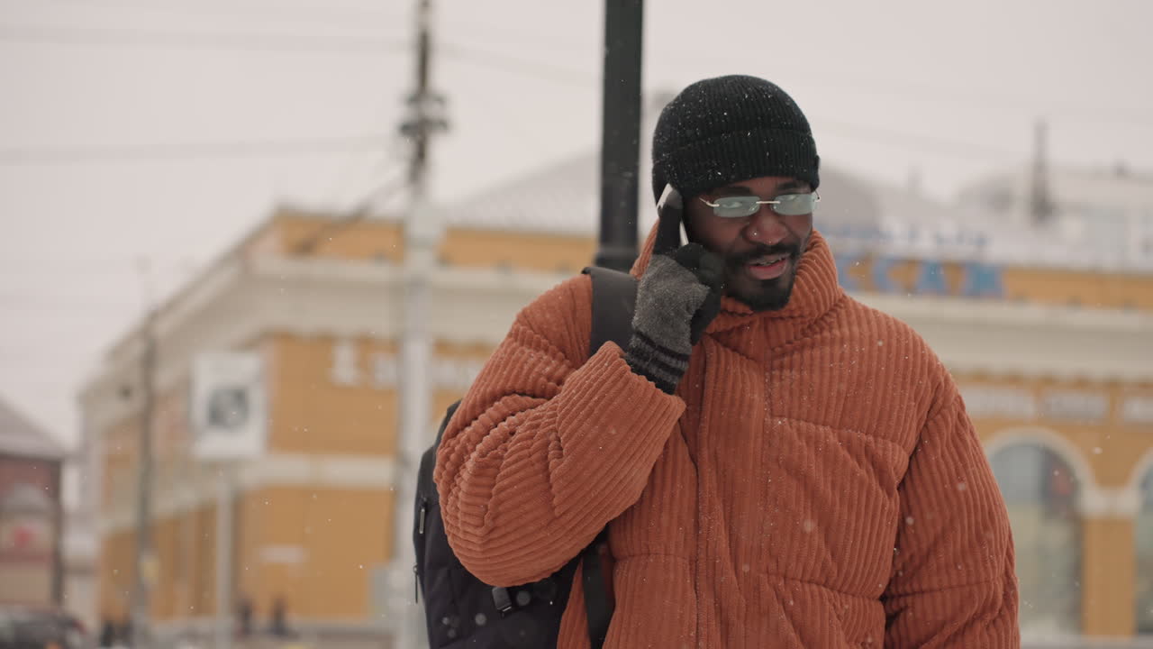 Uomo di colore che parla al telefono in una strada innevata, indossa un cappotto testurizzato color ruggine, berretto nero, occhiali e guanti, zaino a tracolla, fiocchi di neve che cadono delicatamente, espressione concentrata, freelancer che sta contattando.