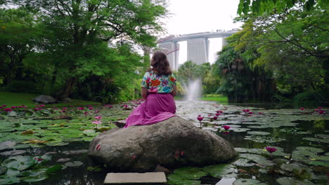 a tourist sit on the rock over water lily pond in gardens by the bay with the supertree grove and marina bay sands backdrop