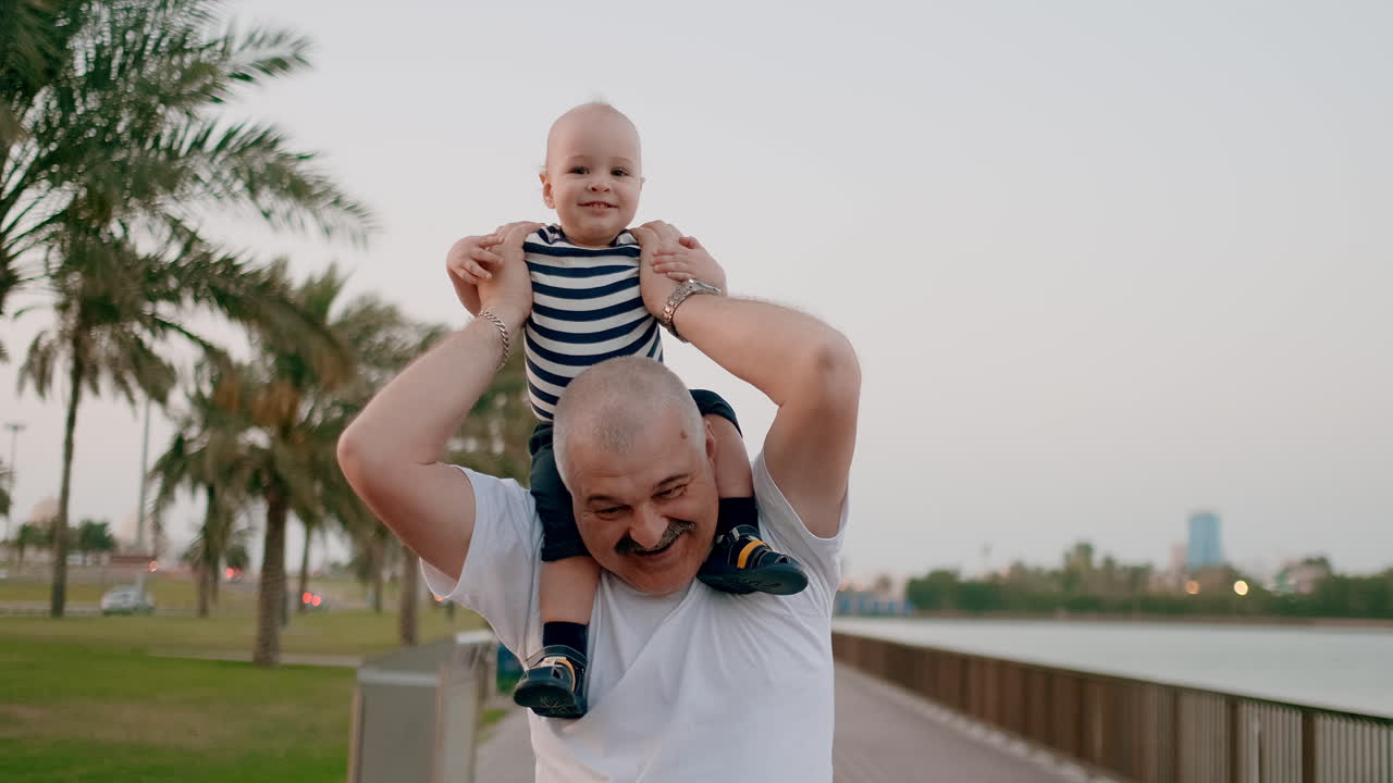 Senior grandpa in a white t-shirt and bristles hipster walking with his son sitting on the neck on the waterfront in the background of the modern city in the summer on the waterfront