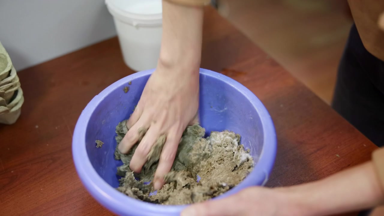 Donna caucasica che mescola la polpa di carta in una ciotola, primo piano di mani che impastano fibre riciclate in una bacinella blu, lavorazione artigianale intima.