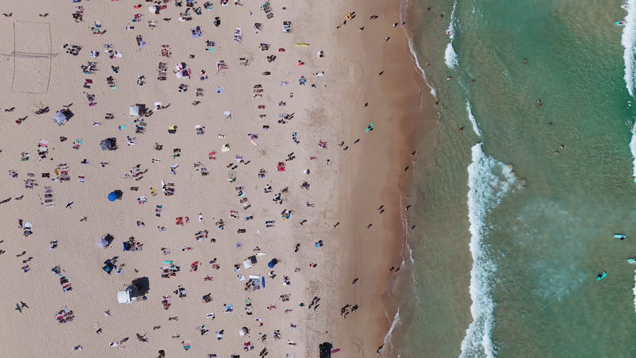 Drone view of Bondi Beach with crowds soaking up the sun and taking dips in the crystal-clear waters. Downward angle aerial shot, New South Wales.