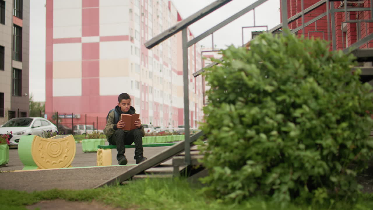 빨간 벽돌 건물 근처 초록색 벤치에 앉아 책을 읽으며 페이지를 넘기는 소년, 어깨에는 백팩을 메고 있고, 식물은 바람에 흔들리고, 흐릿하게 보이는 사람들이 멀리 걸어가는 모습이 보이는 배경이에요.