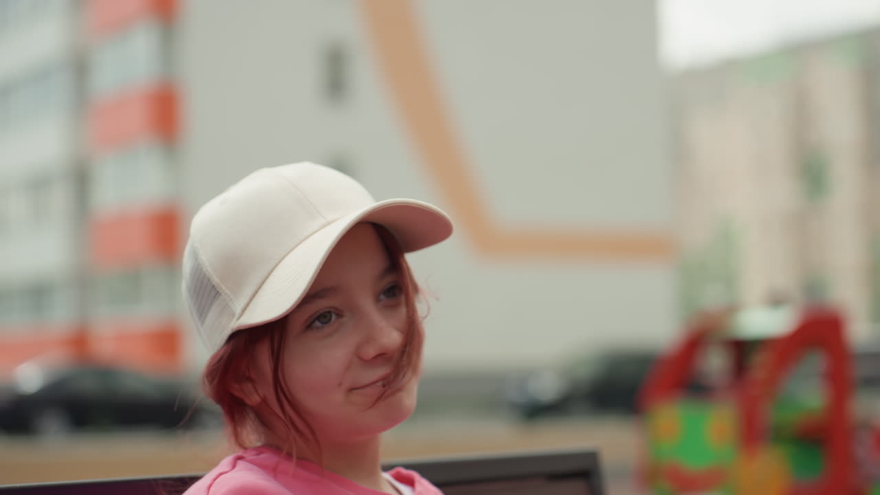 Confident Teenager Sitting On Bench With City Murals And Playground In Background, Cap Tilted And Soft Smile, Relaxed Shoulder And Direct Gaze, Casual Pink Sweater, Urban Lifestyle And Approachable