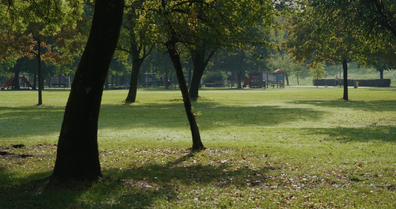 Wide view of a public park in Zagreb, Croatia, with green grass and tall trees casting shadows. Playground structures and benches are visible in the background on a sunny morning.