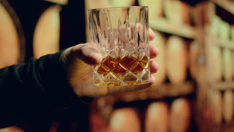 looking carefully at bourbon- slow motion hand swirling glass of bourbon whiskey inside of still house with whiskey barrels in background