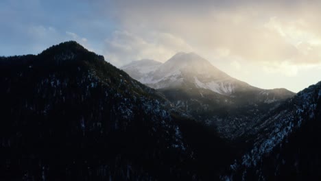 Toma-Aérea-Giratoria-De-Un-Paisaje-Invernal-Del-Monte-Timpanogos-En-El-Fondo-Rodeado-Por-Un-Bosque-De-Pinos-Durante-La-Puesta-De-Sol-Desde-El-Embalse-De-Tibble-Fork-En-El-Cañón-De-La-Horquilla-Americana,-Utah