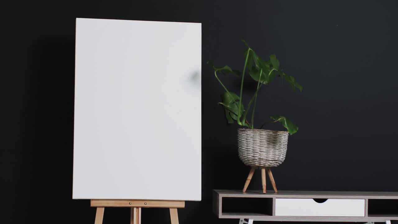 Video of white canvas sign on wooden easel with copy space and plant on desk on black background