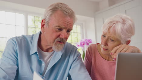 worried retired senior couple with laptop looking at bills at home concerned about cost of living