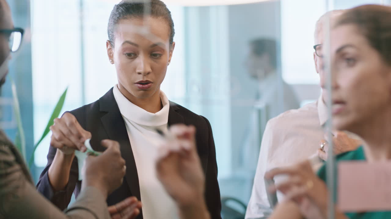 유리 화이트보드를 통해 접착형 메모를 사용하는 사업가들 브레인스토밍 팀 리더 여성 동료들과 함께 사무실 회의에서 문제 해결 전략을 보여줍니다.