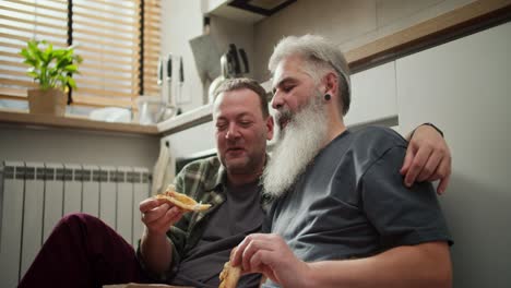 happy elderly man with gray hair and a lush gray beard eats pizza with his boyfriend to a middle-aged brunette with stubble in a green checkered shirt sitting on the floor in the kitchen during lunch