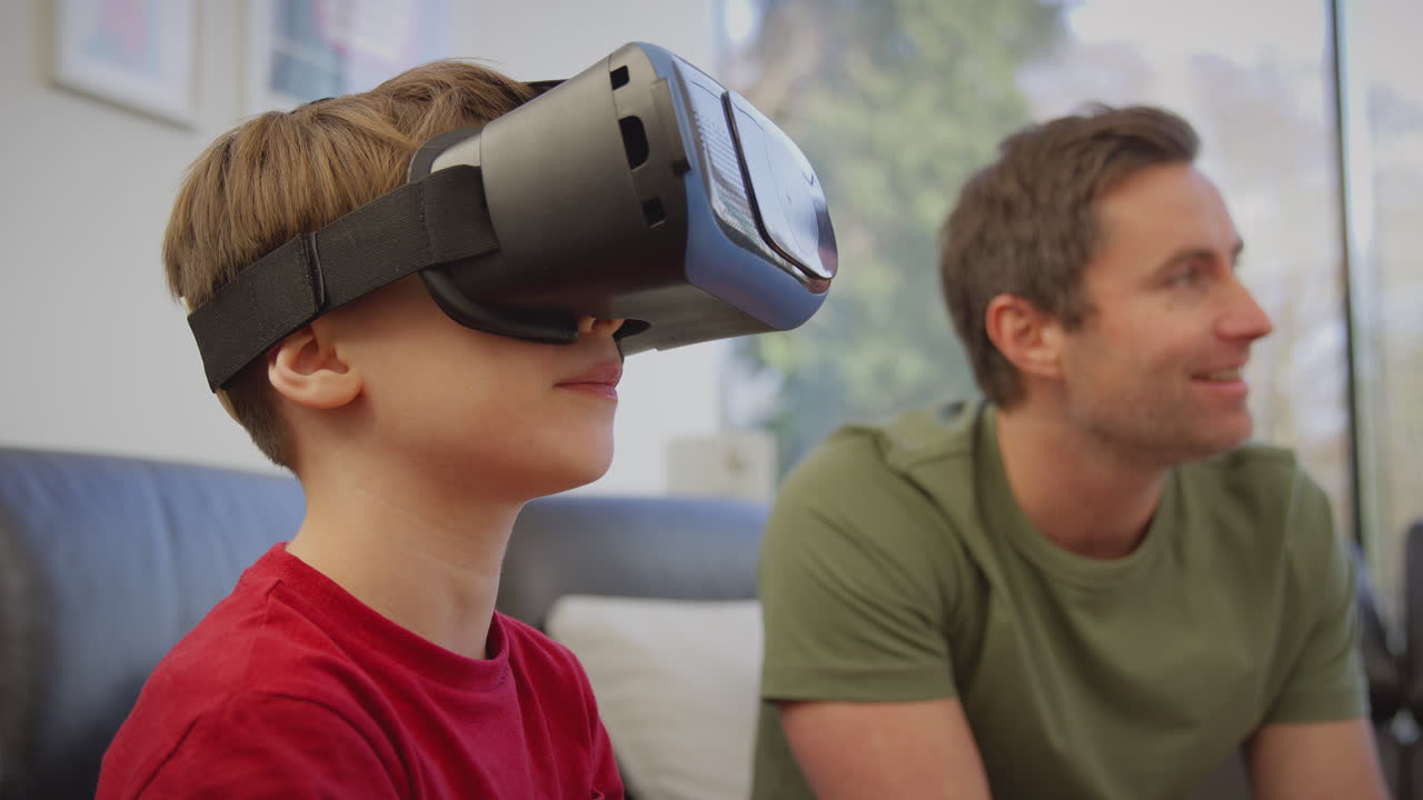 padre viendo a su hijo usando auriculares de realidad virtual jugando a un juego de computadora sentado en el sofá en casa - filmado en cámara lenta
