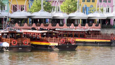 a traditional riverboat glides along a lively urban waterfront, passing colorful riverside buildings and outdoor dining areas in bright daylight with steady camera movement