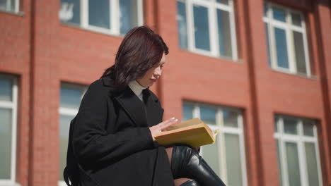 student in black coat and leather boots sitting outside, concentrating on reading her book while flipping to a new page, hair fluttering in the wind with red brick building in background