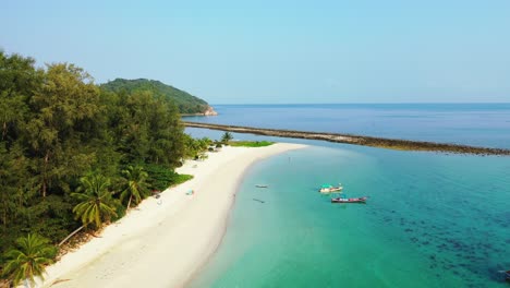 fishing boats anchoring on shore of tropical island with wide sandy beach under palm trees in front of calm turquoise lagoon in thailand