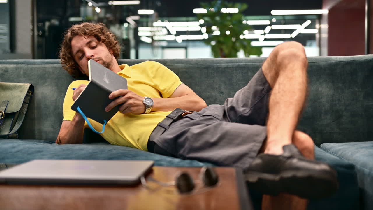 Man in yellow shirt lying on a couch and writing in a notebook