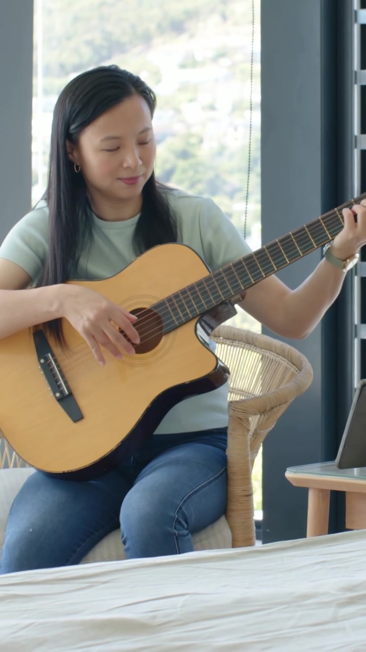 Video vertical: Mujer con camiseta verde y jeans, mirando una tablet y practicando acordes de guitarra en casa.