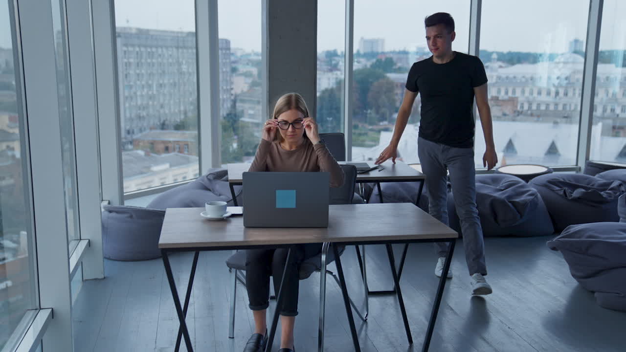 Big spacious modern office with panoramic windows and cityscape view. People sitting at desks working at laptops, male comes up to female to help.