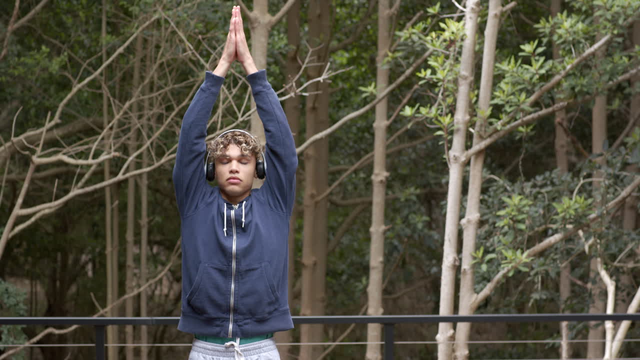 Young man practicing yoga outdoors, wearing headphones, surrounded by trees