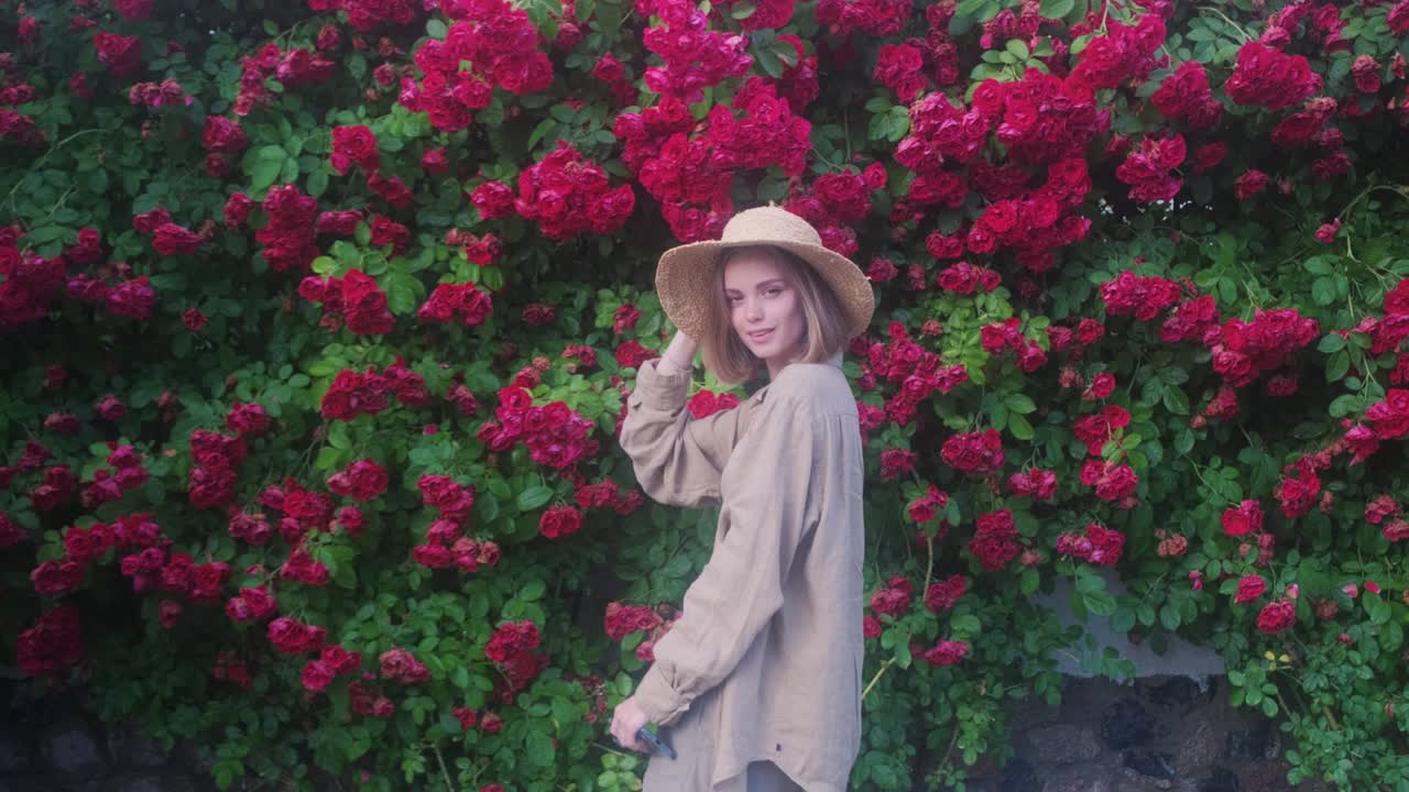 꽃이 피는 장미 덤불의 배경에  모자를 입은 우아한 금발 젊은 여성