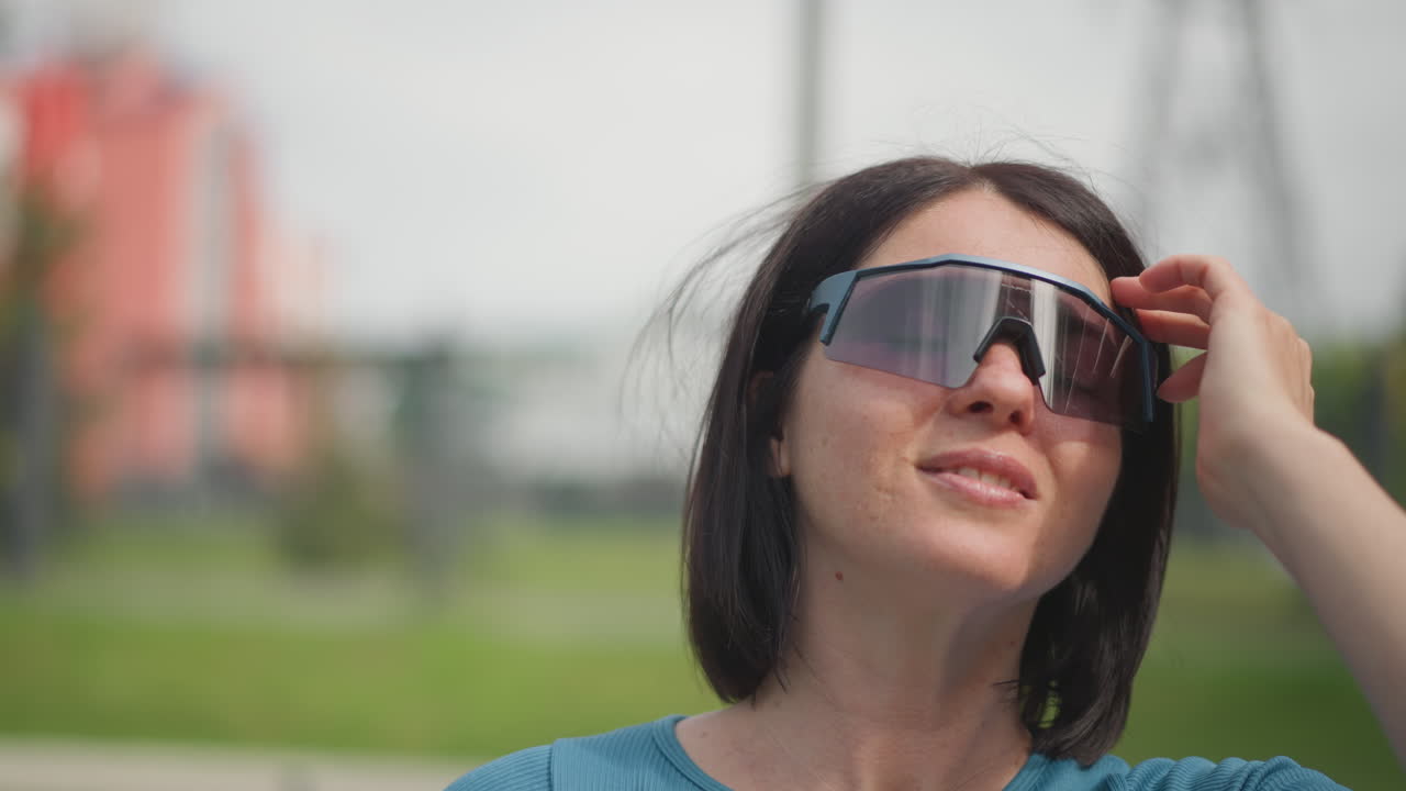 Donna che indossa casco e occhiali da sole in un parco, scena illuminata dal sole che mostra un taglio di capelli corto, visiera riflettente, regolazione del cinturino, espressione sorridente, maglietta casual color teal, preparazione per il tragitto urbano, sicurezza