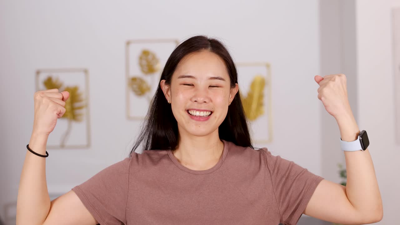 Asian woman smiles, flexes arms in strength pose, brightly lit home, cheerful confident mood