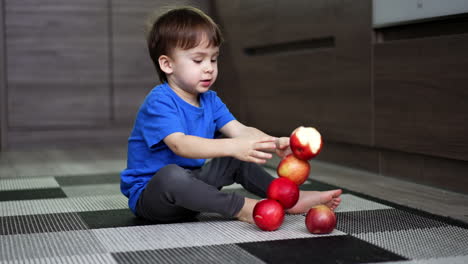 adorable caucasian toddler building a tower of red apples. kid sits on the floor watching the apples fall and roll to the sides.