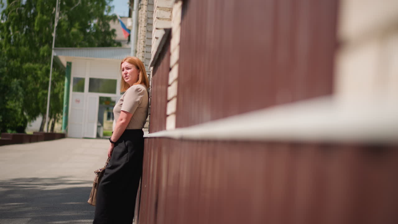 Woman leaning against building wall outdoors under warm summer sunlight, holding bag and gazing thoughtfully into distance, showing calm reflection, emotional fatigue