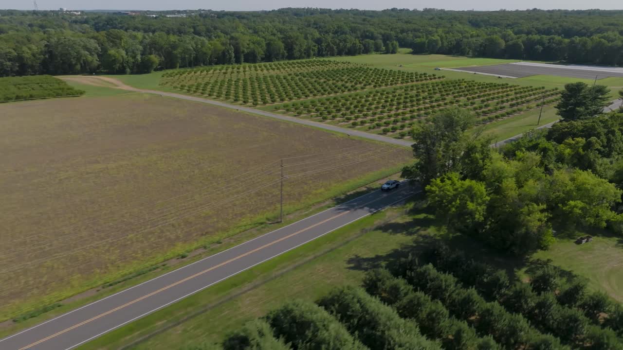 A drone follows a car driving along a picturesque country road surrounded by farm fields and woods