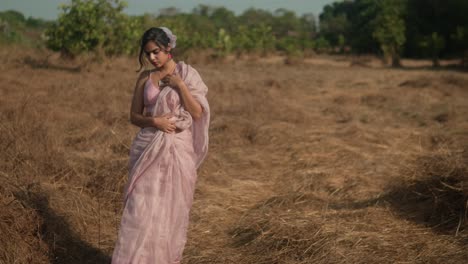 woman in pink saree standing thoughtfully in a dry grass field, trees in background, daytime