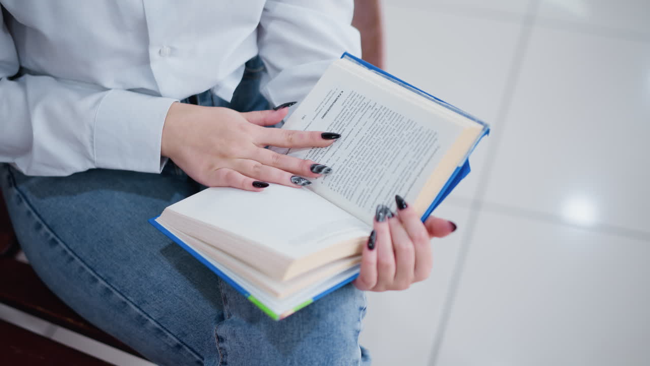 검은색 은 손을 입은 청바지 바지를 입은 여성이 나무 벤치에 앉아 밝은 쇼핑몰에서 책 페이지를 뒤집고 부드러운 타일이 빛을 반사합니다.