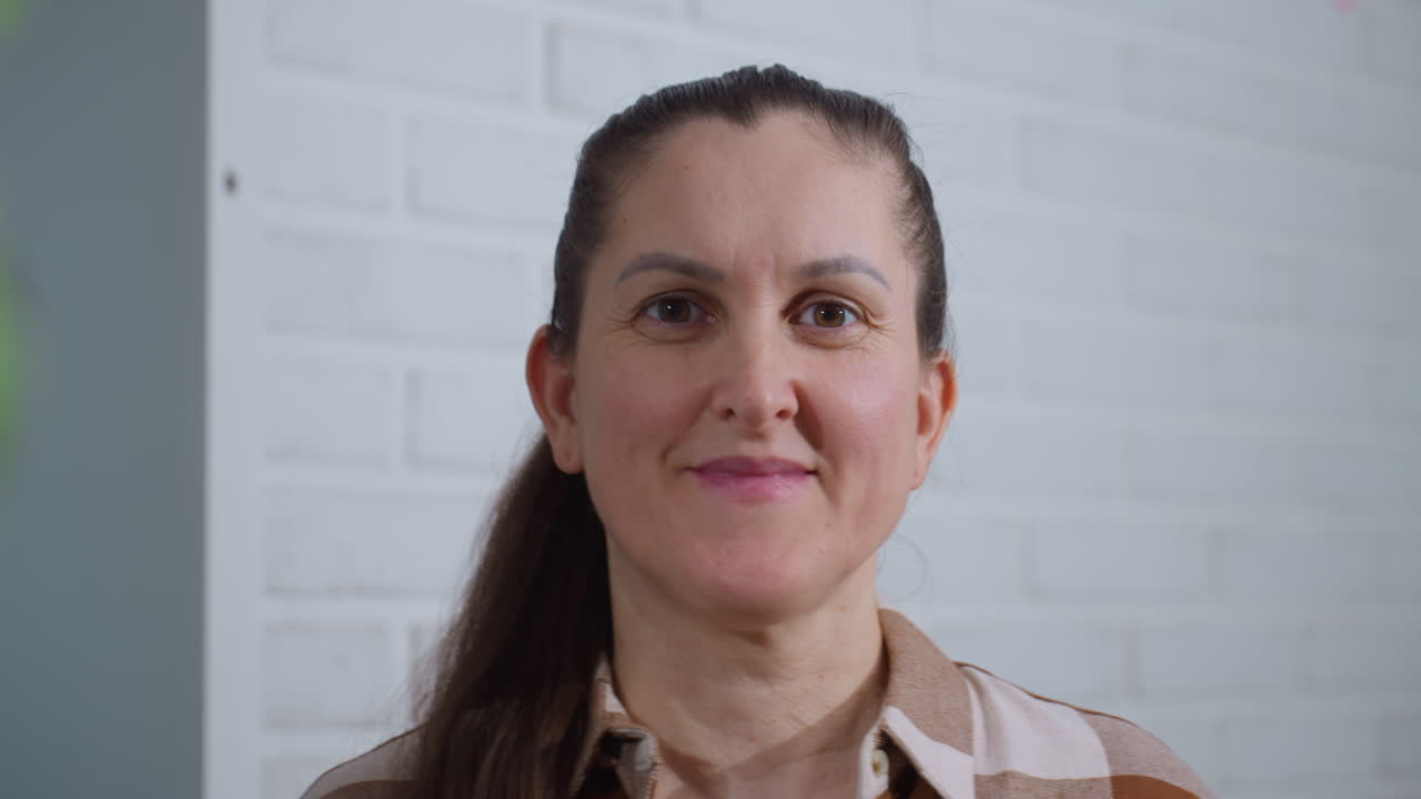 Elegant lady with bright smile wearing plaid shirt stands before white brick wall, subtle green plant blur at side, warm natural light accentuating friendly expression and relaxed indoor atmosphere